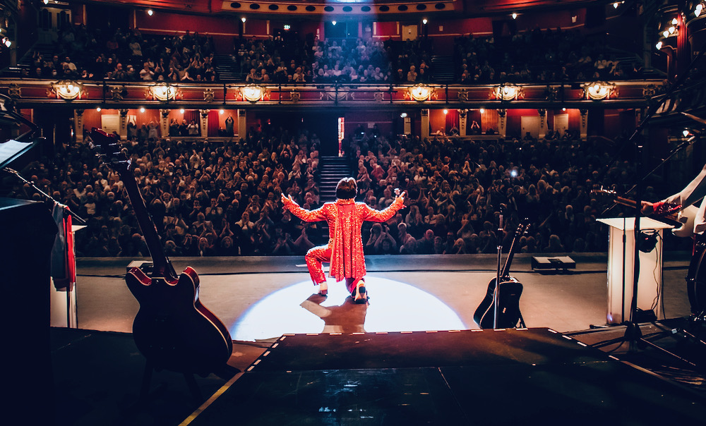 Elvis tribute performer in an owl jumpsuit kneeling on stage before a sold-out theatre audience during a live show