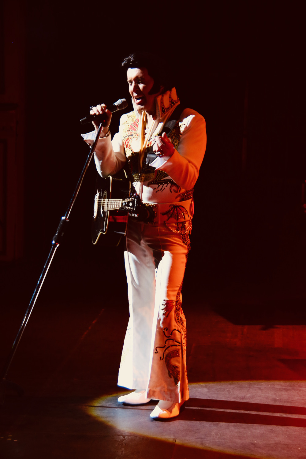 Elvis tribute performer in a dragon jumpsuit holding a guitar at a microphone during a live show