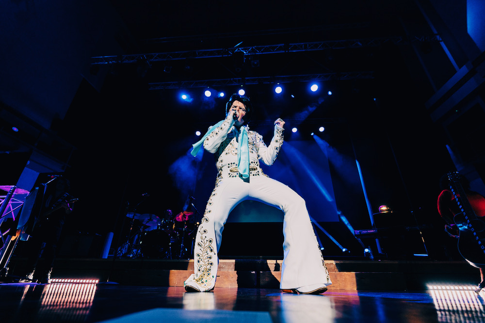 Rob Kingsley performing A Vision of Elvis live on stage in the White Spanish Flower Elvis jumpsuit under blue concert lights